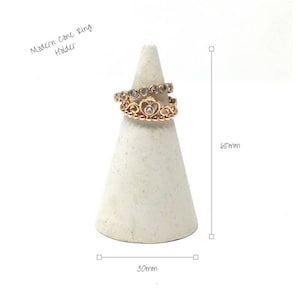 May include: A white cone ring holder with a gold ring with small clear stones and a heart design. The holder is 6.5 inches tall and 3 inches wide. "Modern Cone Ring Holder" is written above the holder.