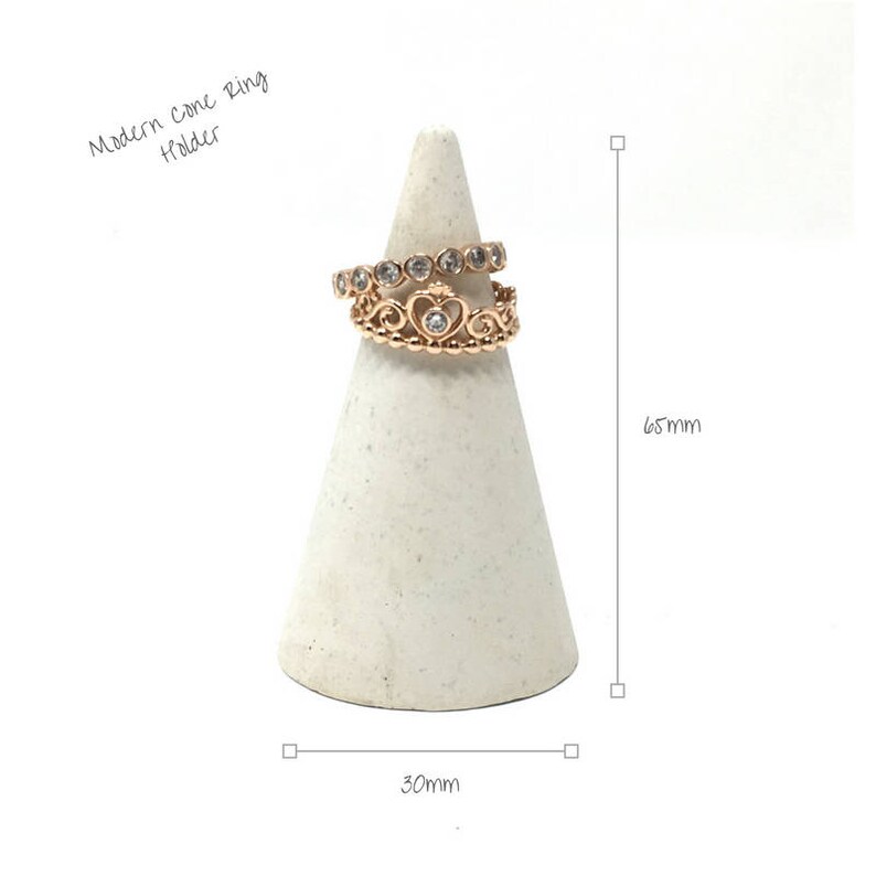 May include: A white cone ring holder with a gold ring with small clear stones and a heart design. The holder is 6.5 inches tall and 3 inches wide. "Modern Cone Ring Holder" is written above the holder.