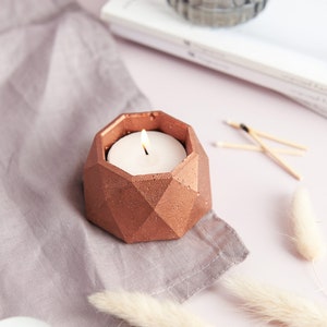 Peut inclure: Un bougeoir géométrique de couleur cuivre avec une bougie chauffe-plat blanche à l'intérieur. La bougie est allumée et la flamme est visible. Le bougeoir est posé sur une surface en tissu gris.