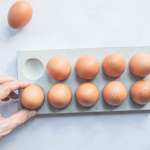 Puede incluir: Un porta huevos de hormig&oacute;n gris con diez huecos, nueve de los cuales est&aacute;n llenos de huevos marrones. Una mano est&aacute; colocando un huevo marr&oacute;n en el hueco vac&iacute;o.