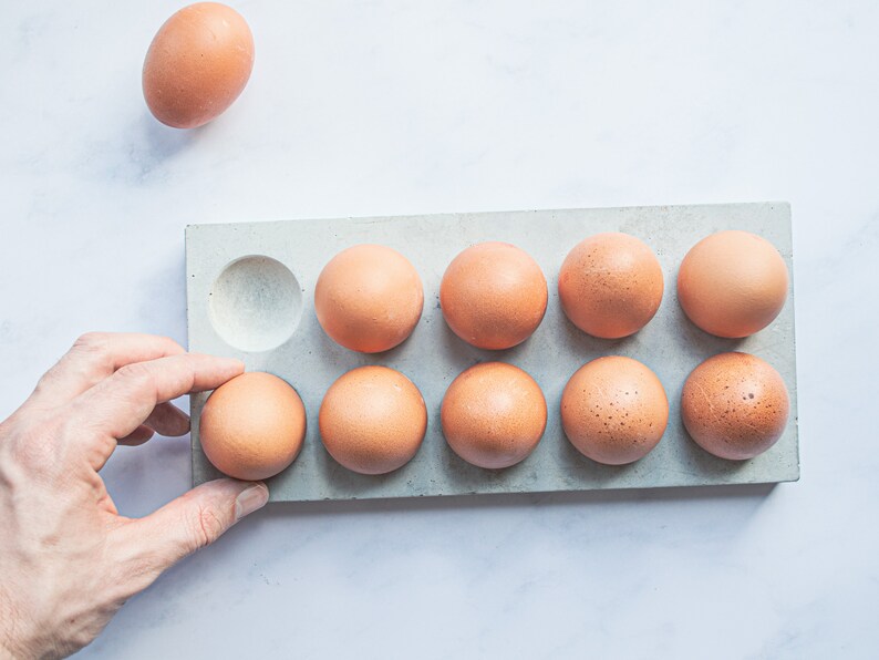 Puede incluir: Un porta huevos de hormig&oacute;n gris con diez huecos, nueve de los cuales est&aacute;n llenos de huevos marrones. Una mano est&aacute; colocando un huevo marr&oacute;n en el hueco vac&iacute;o.