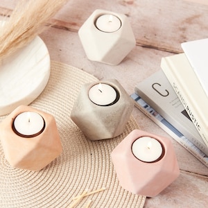 Op de afbeelding: Vier geometrische kaarshouders in verschillende kleuren: beige, grijs, terracotta en roze. Elke houder heeft een kleine, ronde opening voor een theelichtje.