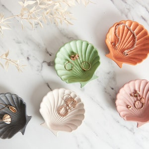 May include: Five decorative seashell-shaped dishes in different colours, including green, white, pink, grey, and orange. Each dish is filled with jewellery, such as rings, earrings, and necklaces.