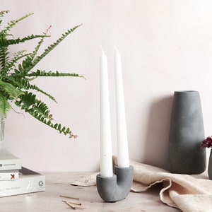 May include: A grey concrete candle holder with two white candles. The holder is shaped like a U and has a rustic look. There are two books, a potted fern, and a grey vase in the background.