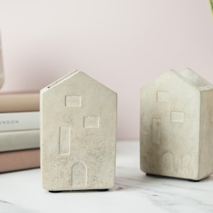 Könnte beinhalten: Zwei dekorative Objekte in Form eines Betonhauses. Jedes hat einen rechteckigen Körper mit einem Satteldach und Tür- und Fensterausschnitten. Die Objekte sind hellgrau und stehen auf einer weißen Oberfläche. Eine rosa-weiß gestreifte Vase und Bücher befinden sich im Hintergrund.