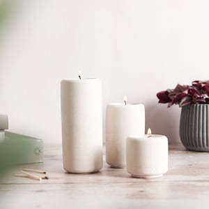Op de afbeelding: Drie witte betonnen kaarsen in verschillende maten, waarbij de grootste kaars brandt en de twee kleinere kaarsen ook branden. De kaarsen staan op een licht houten oppervlak met een plant in een pot en boeken op de achtergrond.