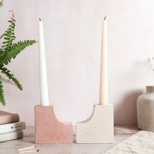 May include: Two concrete candle holders, one pink and one gray, with tall white and cream candles. The holders are on a wooden surface with a white and gray fern plant in a white pot and a gray vase with dried white flowers.