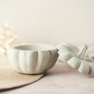 Puede incluir: Un recipiente gris claro en forma de calabaza con una tapa a juego. El recipiente está abierto, revelando el interior. La tapa está ligeramente entreabierta, mostrando el detalle del tallo. El recipiente se encuentra sobre una estera tejida, con flores secas en el fondo.