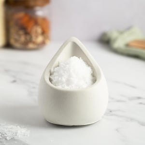 Puede incluir: Un pequeño salero de cerámica blanca lleno de sal gruesa blanca. El recipiente en forma de lágrima se encuentra sobre una superficie de mármol blanco. Otros frascos y una cuchara de madera son visibles en el fondo.