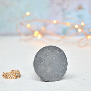 May include: A gray concrete disc with a rough texture sits on a white surface. A gold ring with a crown design and a clear stone is in the foreground.
