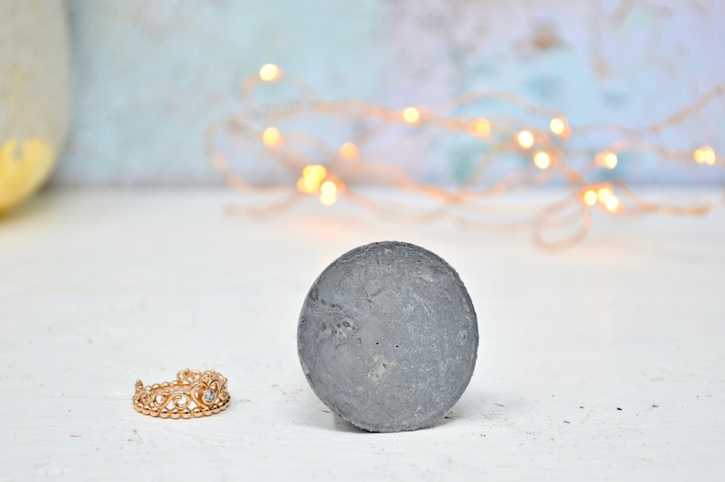 May include: A gray concrete disc with a rough texture sits on a white surface. A gold ring with a crown design and a clear stone is in the foreground.