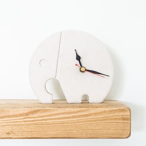 Può includere: Orologio a forma di elefante in ceramica bianca con lancette nere e una lancetta dei secondi rossa. L'orologio è appoggiato su uno scaffale di legno.
