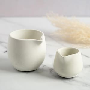 May include: Two off-white ceramic pitchers with a matte finish. The larger pitcher is taller and wider than the smaller one. Both have a spout for pouring. The background includes a marble surface and dried ornamental grass.
