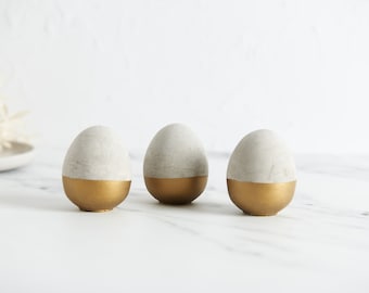 May include: Three decorative concrete eggs painted with a gold band around the bottom half. The eggs are arranged in a row on a white marble surface.