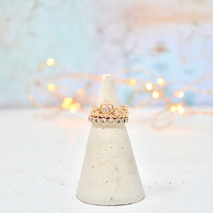 May include: A gold ring set with a heart-shaped stone, surrounded by smaller stones, on a white cone-shaped ring holder. The ring is on a white surface with a blurred background of white and blue with string lights.