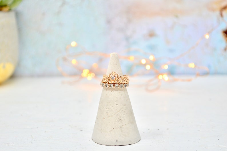 May include: A gold ring set with a heart-shaped stone, surrounded by smaller stones, on a white cone-shaped ring holder. The ring is on a white surface with a blurred background of white and blue with string lights.