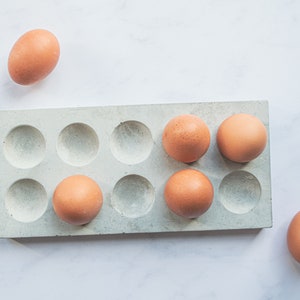 Puede incluir: Un porta huevos de hormig&oacute;n blanco con seis huevos marrones dentro y un huevo marr&oacute;n fuera del porta huevos.