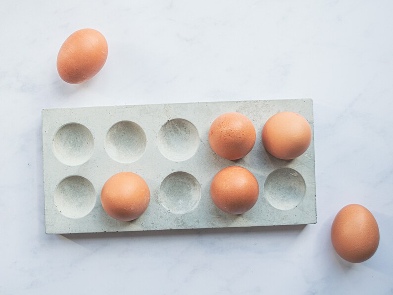 Puede incluir: Un porta huevos de hormig&oacute;n blanco con seis huevos marrones dentro y un huevo marr&oacute;n fuera del porta huevos.