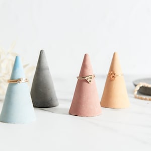 May include: Four small concrete cone-shaped ring holders in blue, gray, pink, and beige. Each holder has a gold ring on top.