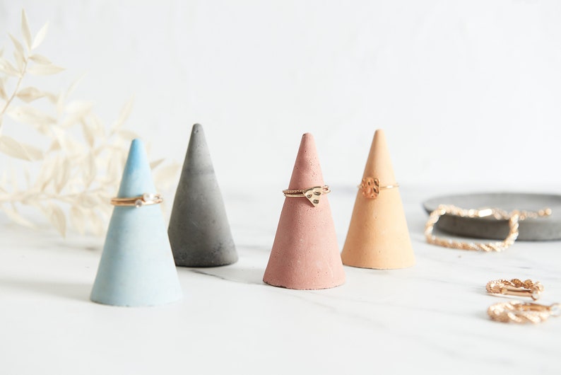 May include: Four small concrete cone-shaped ring holders in blue, gray, pink, and beige. Each holder has a gold ring on top.