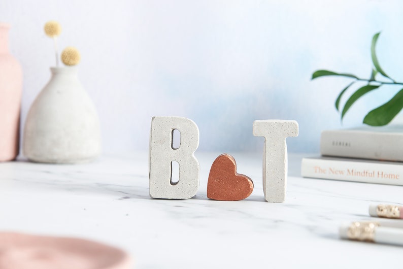K&ouml;nnte beinhalten: Wei&szlig;e Betonbuchstaben "B" und "T" mit einem kleinen Kupferherz dazwischen. Die Buchstaben stehen auf einer wei&szlig;en Marmorfl&auml;che.