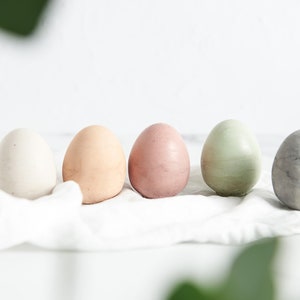 Può includere: Cinque uova di Pasqua dipinte in tonalità pastello disposte in fila su un panno di lino bianco. Le uova sono dipinte in tonalità di bianco, pesca, rosa, verde e grigio.