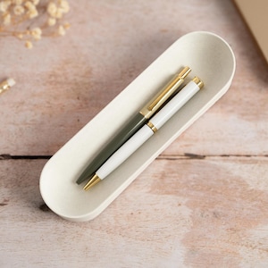May include: A cream-coloured, oblong tray contains two pens: one dark green and gold, the other white and gold. The pens are on a light wooden surface, with two pencils and dried flowers in the background. The scene is well-lit, suggesting a workspace.