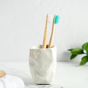 Puede incluir: Un portacepillos de cerámica blanca texturizada con dos cepillos de dientes de bambú. Uno tiene cerdas blancas y el otro cerdas turquesas. Una pastilla de jabón y una toalla blanca doblada están en primer plano.