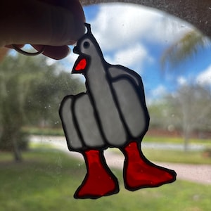 May include: A stained glass pigeon keychain. The bird is gray with red feet and a red beak. The keychain is held up in front of a window with a view of the sky and trees.
