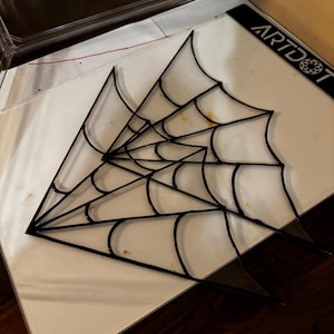 May include: A black spiderweb design on a white surface. The web is made of black lines and is partially transparent. The word "ARTDOT" is visible on the right side of the image.