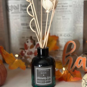 May include: A black glass bottle with a green label that reads "Shower N Shave" and "Idlewylde Fire Reed Diffuser" with a white flower and wooden sticks. The bottle is sitting on a white surface with fall leaves and pumpkins in the background.