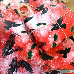 May include: Red wrapping paper with a pattern of black crows, holly, and white speckles. A wrapped gift is tied with twine. Scissors and a ball of twine are also visible.