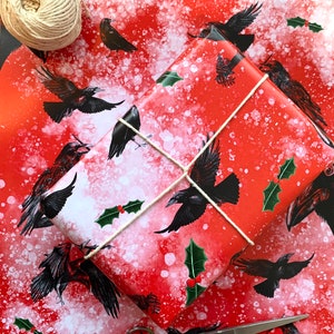 May include: Red and white patterned wrapping paper with black raven illustrations and holly accents. A wrapped gift is tied with twine, and a ball of twine and scissors are nearby. A festive theme.