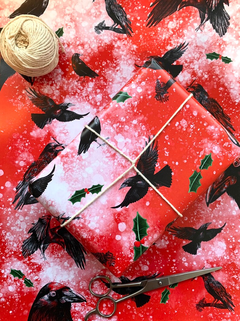 May include: Red and white patterned wrapping paper with black raven illustrations and holly accents. A wrapped gift is tied with twine, and a ball of twine and scissors are nearby. A festive theme.