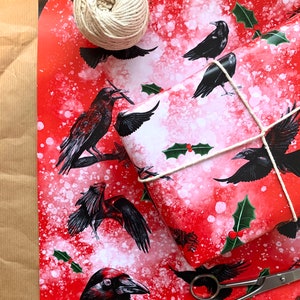 May include: Red and white patterned wrapping paper with black raven illustrations, holly sprigs, and white speckles. A roll of twine, wrapped gift, and scissors are also present. The paper is on a brown paper surface.