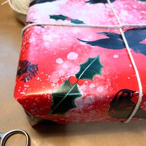 May include: A wrapped gift with red wrapping paper featuring black ravens and holly leaves. The gift is tied with twine and sits on a brown paper surface. A ball of twine and scissors are also visible.