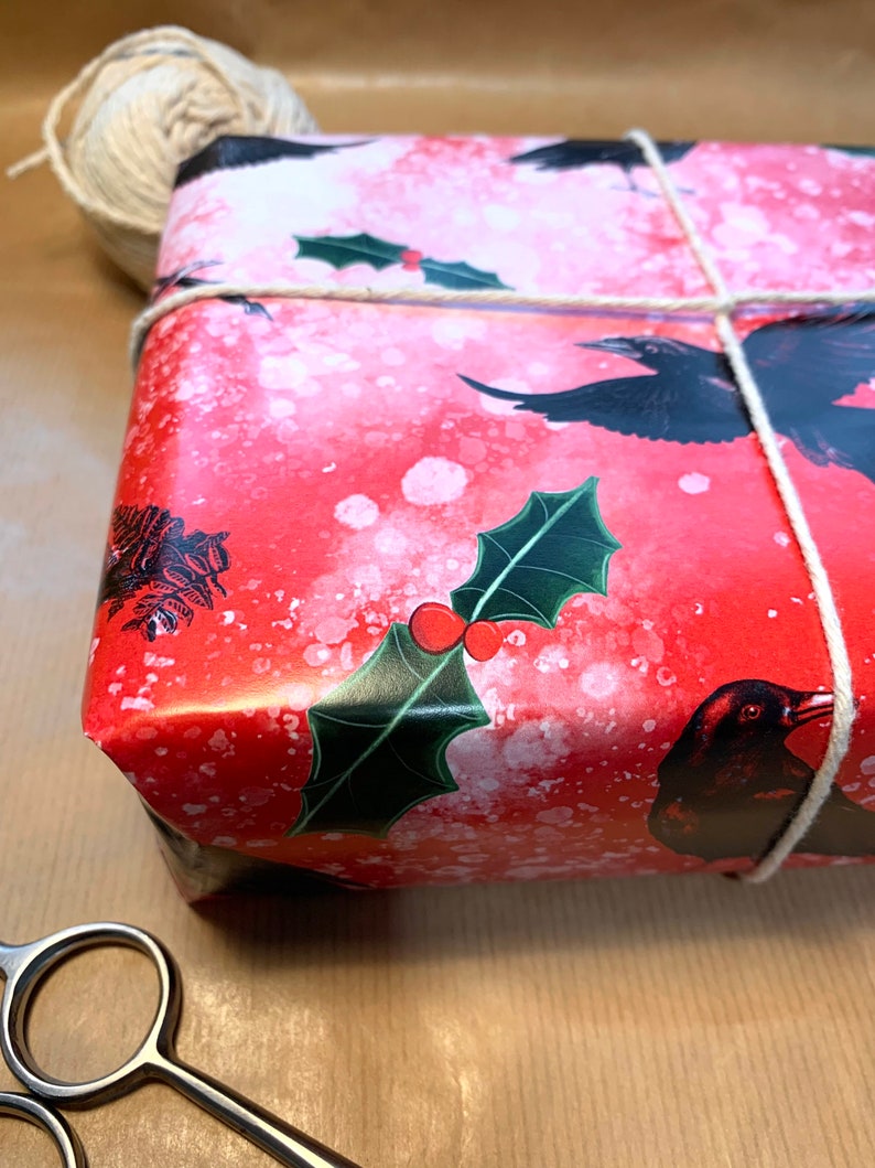 May include: A wrapped gift with red wrapping paper featuring black ravens and holly leaves. The gift is tied with twine and sits on a brown paper surface. A ball of twine and scissors are also visible.