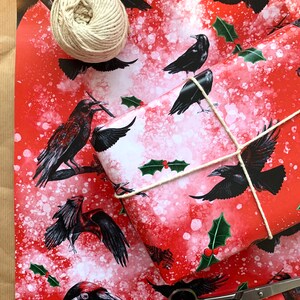 May include: Red wrapping paper with a pattern of black crows and holly leaves. A wrapped gift is tied with twine, and a ball of twine and scissors are nearby. The paper has a white speckled background.