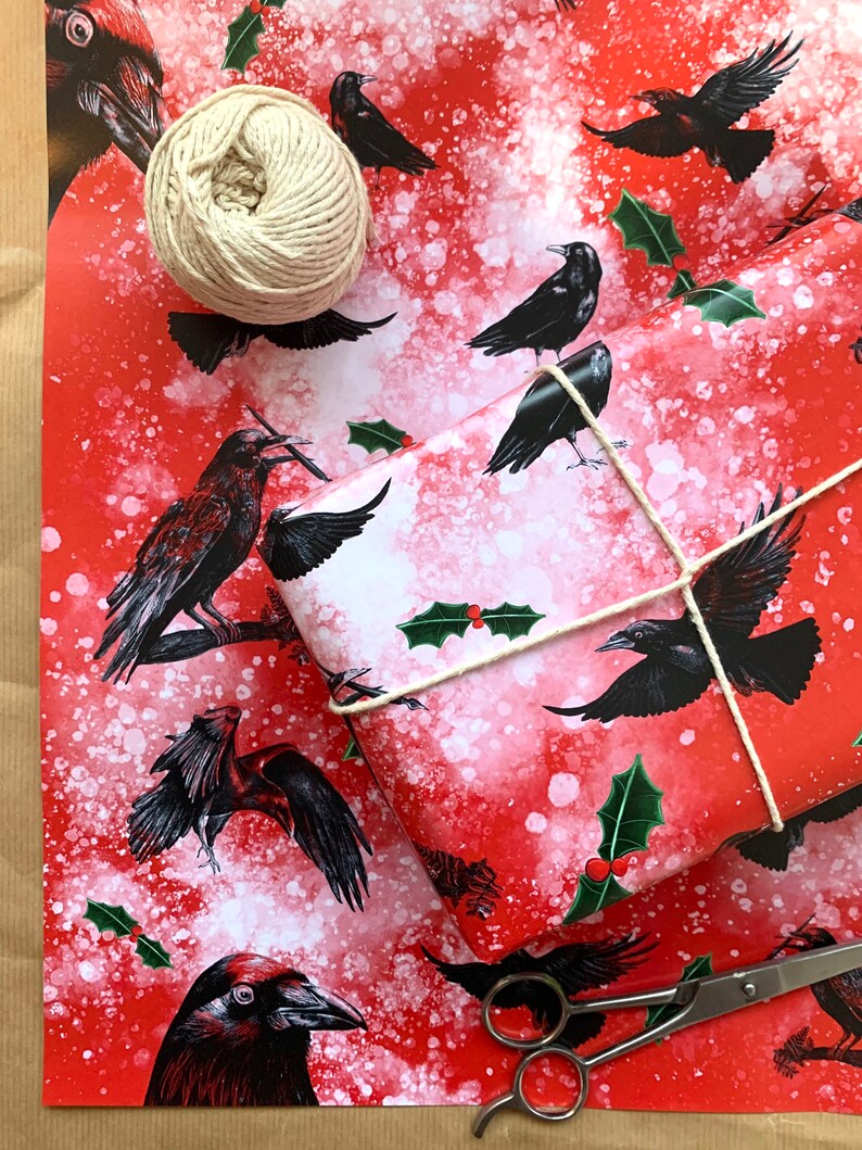 May include: Red wrapping paper with a pattern of black crows and holly leaves. A wrapped gift is tied with twine, and a ball of twine and scissors are nearby. The paper has a white speckled background.