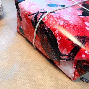 May include: A wrapped gift with red wrapping paper featuring black raven illustrations and white speckles. The gift is tied with natural twine. The background is a light brown surface.