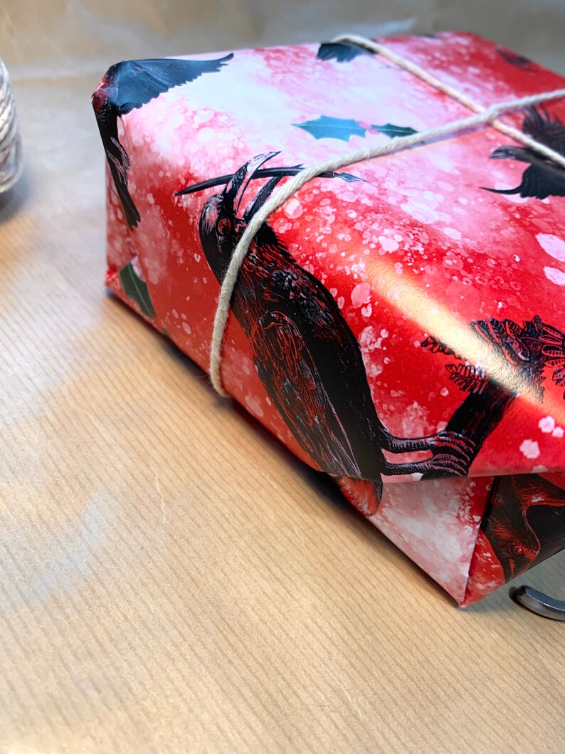 May include: A wrapped gift with red wrapping paper featuring black raven illustrations and white speckles. The gift is tied with natural twine. The background is a light brown surface.