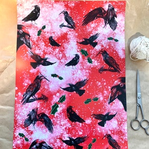 May include: Red wrapping paper with a pattern of black crows in various poses, with sprigs of holly and white accents. A ball of white twine and silver scissors are next to the paper.