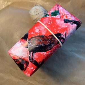 May include: A wrapped gift with red wrapping paper featuring a black bird design and white speckles. The gift is tied with twine and rests on a brown paper surface. A ball of twine and scissors are also present.