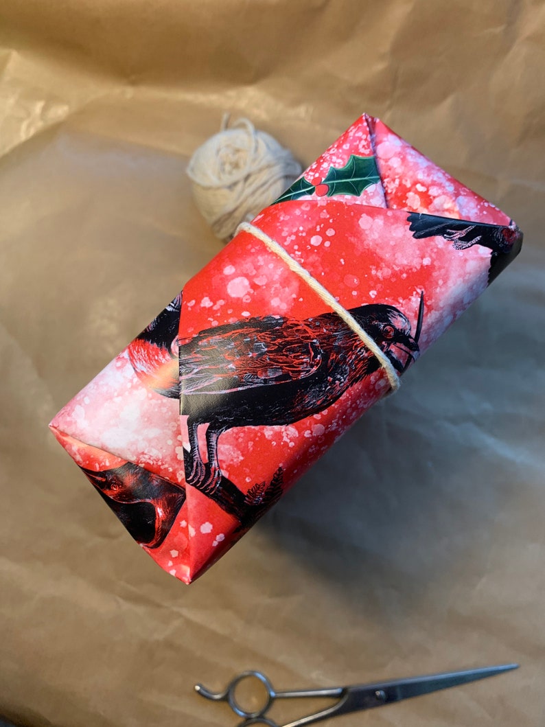May include: A wrapped gift with red wrapping paper featuring a black bird design and white speckles. The gift is tied with twine and rests on a brown paper surface. A ball of twine and scissors are also present.