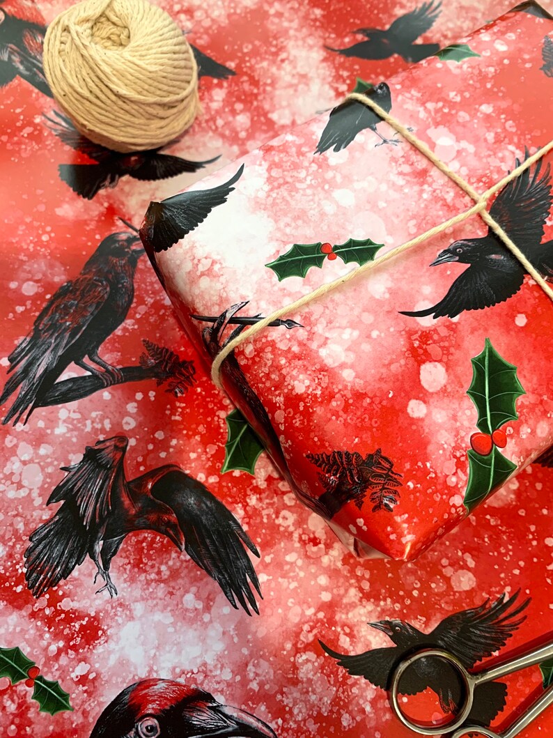 May include: Red wrapping paper with black raven illustrations and white speckles. A wrapped gift is tied with twine and decorated with holly. A ball of twine and scissors are also visible.
