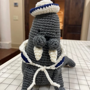 May include: A gray crocheted walrus wearing a white and blue sailor hat and a white and blue sailor scarf. The walrus has white crocheted tusks.