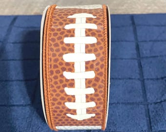 May include: A brown and white ribbon with a football design. The ribbon has a brown background with white stitching and a white football pattern.