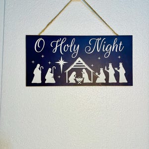 May include: A dark blue wooden sign with white lettering that reads "O Holy Night". The sign features a white silhouette of the nativity scene with a star above the manger.