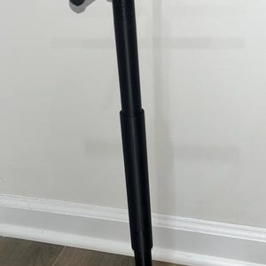 May include: A black, adjustable display stand with a circular base and a T-shaped top. The stand is made of metal and has a matte finish. The top has four horizontal bars for displaying items. The stand is designed to hold and showcase various products.