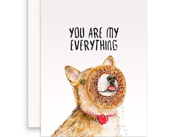 May include: A greeting card featuring a watercolour illustration of a dog with a doughnut over its face. The text on the card reads "You are my everything."
